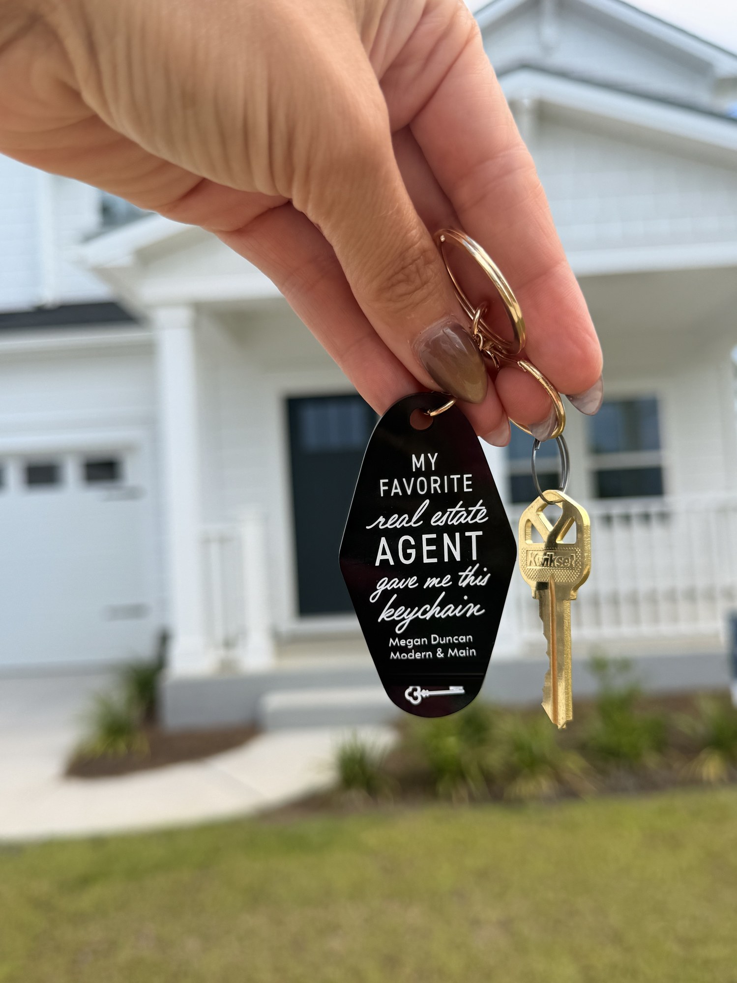 A hand holding a house key and a black keychain that reads: my favorite real estate agent gave me this keychain, Megan Duncan