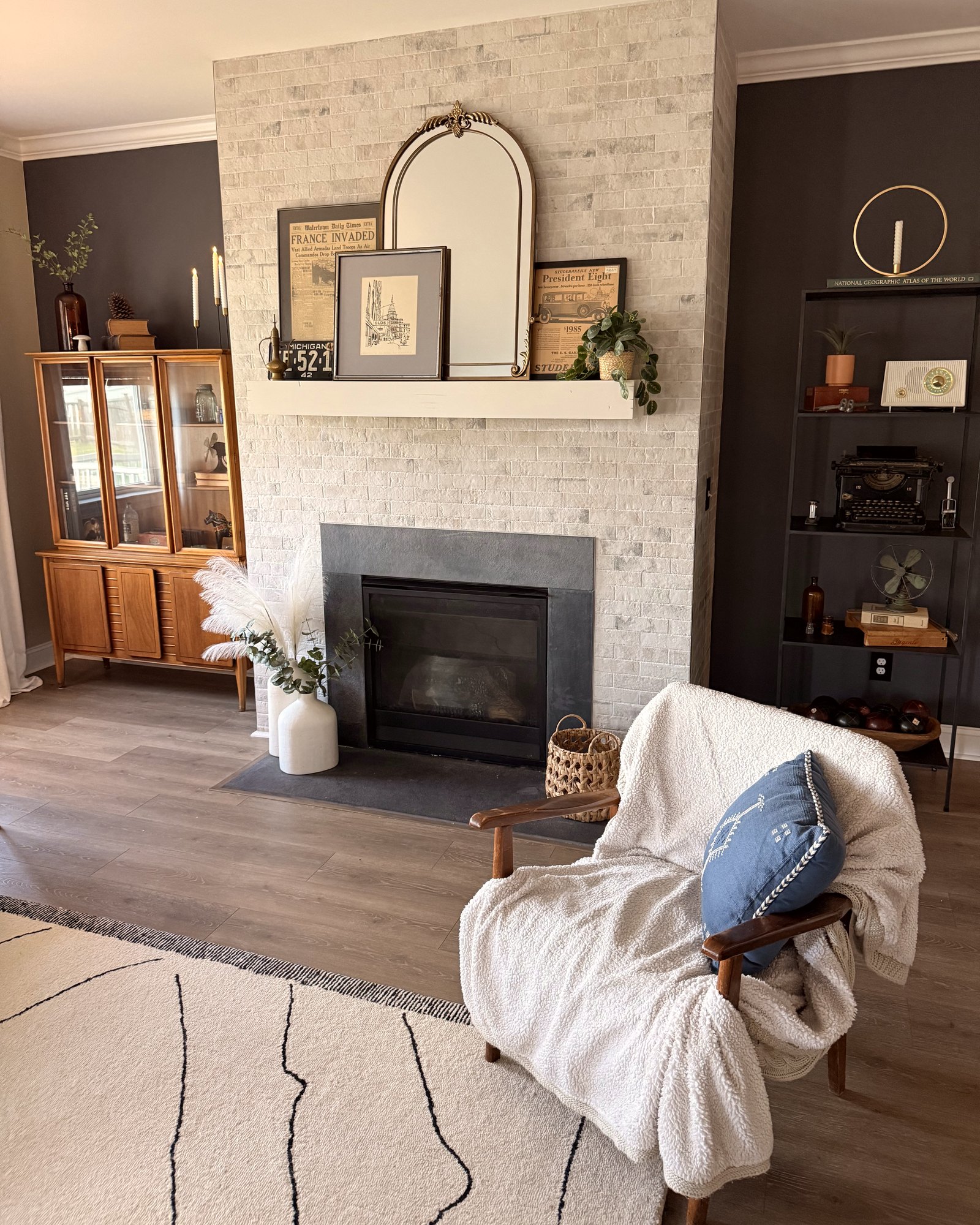 A styled living room with a whitewashed brick fireplace, an arched brass mirror, and a rocking chair draped with a sherpa throw